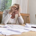 A woman sitting at a desk with her head in her hands, surrounded by paperwork