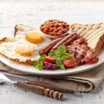 A hearty breakfast plate featuring fried eggs, bacon, sausage, baked beans, toast, and salad