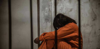 Person in orange jumpsuit sitting behind bars, head down.
