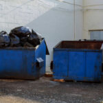 Two blue dumpsters, one overflowing with trash bags.