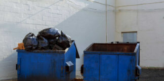 Two blue dumpsters, one overflowing with trash bags.