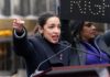 A woman passionately speaking at a political rally with a sign in the background