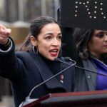 A woman passionately speaking at a political rally with a sign in the background