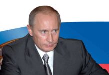 GONE: Putin Vanishes as Russia Bleeds Out A serious-looking man in a suit sitting at a table with a Russian flag in the background