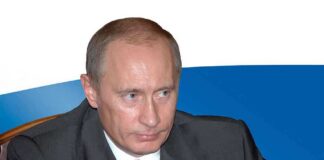 A serious-looking man in a suit sitting at a table with a Russian flag in the background