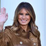 Melania Trump waving and smiling while wearing a stylish coat